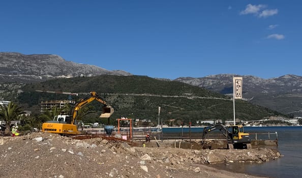 Budva,Montenegro-April 11,2025:A Volvo excavator working on a waterfront construction site with mountains and blue sea in the background. There is a flag with the word COCO next to it. High quality photo