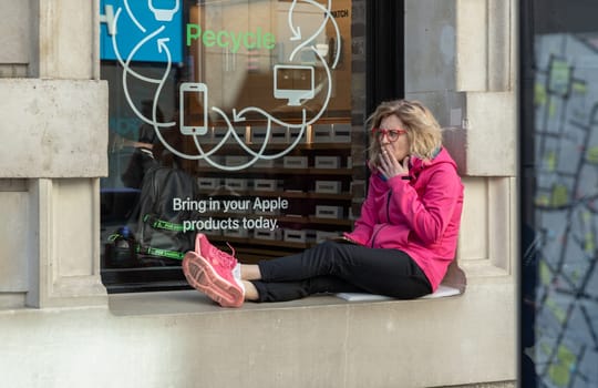 London, UK - May 13, 2025 - A middle-aged woman with blonde hair and red-rimmed glasses is sitting on the glass window frame outside a shop and smoking a cigarette. use it as your Wallpaper, Poster and Space for text, Selective focus.