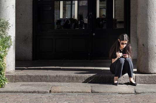 London, UK - May 13, 2025 - A long-haired woman in a black dress and sunglasses sits on the edge of the walkway and looking view messages at her smartphone. use it as your Wallpaper, Poster and Space for text, Selective focus.