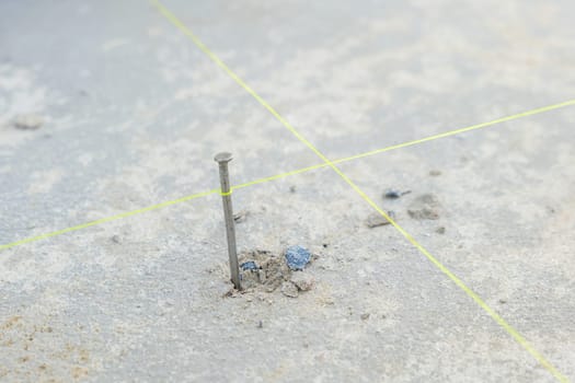 Nail hammered into rough concrete, with bold intersecting yellow lines at its base, highlighting precision for outdoor tiling alignment around the home.