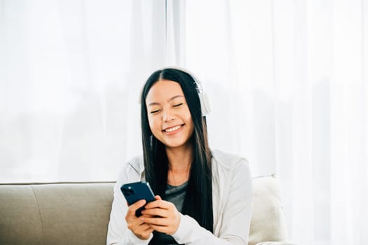 Asian woman on sofa relaxes listens to music watches movies on mobile. Engaged in resting enjoyment and technology for leisure at home.