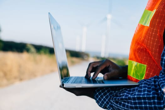 Technician with laptop by windmills an engineer at work. Service technician ensures turbine efficiency. Confident in landscape development focused on windmill service and quality.
