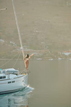 A beautiful girl in a pareo stands on the bow of a sailing boat and waves at sunrise, a sexy figure, mountains in the background, she enjoys the views and azure color of the sea on a sunny day. High quality photo