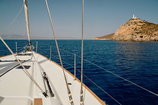 view from a sailing boat to an island with a lighthouse in clear sunny weather, the bow of the boat, the shrouds of the yacht, reflection on the water, mountains in the background. High quality photo