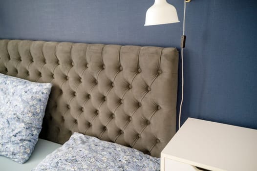 Modern upholstered headboard close-up with floral pillows in cozy bedroom. White pendant light and gray headboard, cozy minimalist bedroom. white nightstand. High quality photo