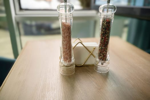 Salt and pepper mills on a wooden table and paper napkins. Pink salt, coarse grind. Black and red pepper.