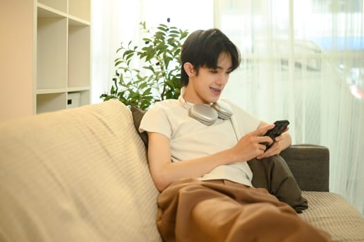 Smiling young man with headphones around his neck lounges on sofa and using smartphone.