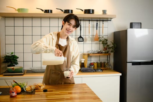 Cheerful young man pours fresh milk into a glass, standing in a modern kitchen.