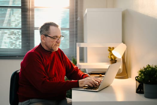 Senior man in red sweater working online in a calm room with natural and lamp light.