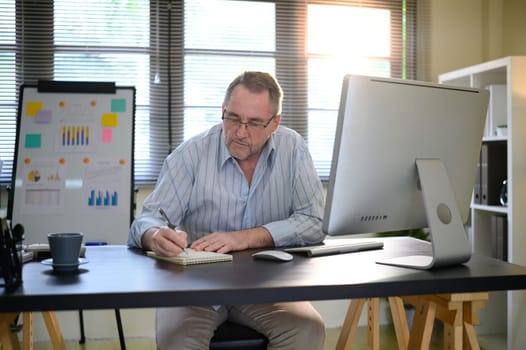Mature businessman sits at workspace, taking handwritten notes while analyzing charts and documents,.