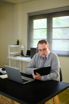 Focused senior man in glasses works at home with digital tablet and laptop.