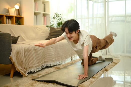Smiling young asian man practices yoga on a mat in his living room.