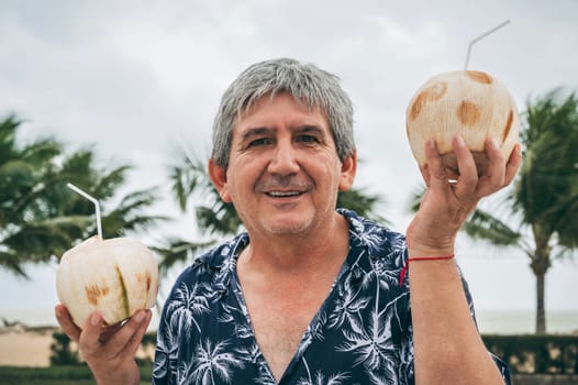 happy old male senior tourist holds a drinking coconut with a straw on vacation on beach by the sea