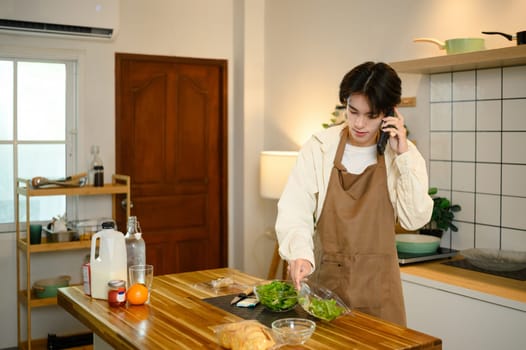 Young man talks on the phone while making a fresh green salad at home.
