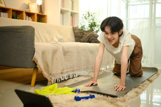 Young man follows an online fitness session while exercising on a mat in his living room.
