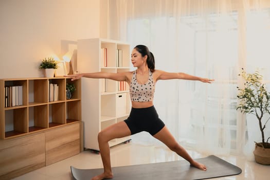 Young woman practices Warrior II yoga pose on mat in a bright living room.