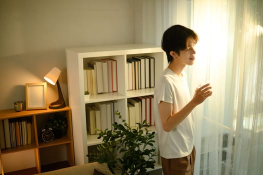 Young man standing by the window, enjoying the morning sunlight in a cozy home.