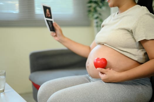 Pregnant woman holding ultrasound and red heart, maternity love and anticipation at home.