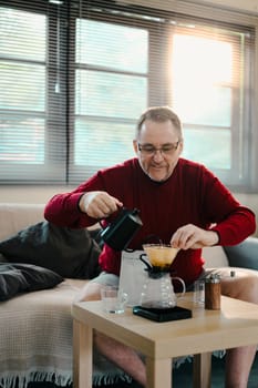 Happy elderly man carefully making pour-over coffee at home with a glass dripper and kettle.