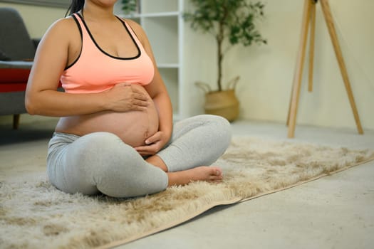 Pregnant woman in activewear sits on the floor with hands resting on her bump.