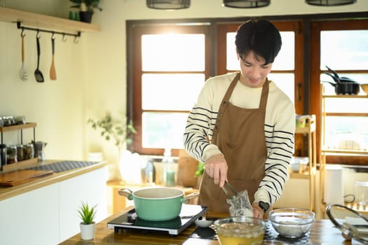 Smiling man in an apron prepares noodles over a stove in warm modern kitchen.