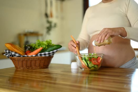 Pregnant woman adds vinaigrette to a colorful salad at a kitchen counter.