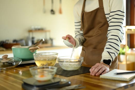 Smiling man in an apron prepares noodles over a stove in warm modern kitchen.