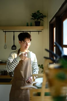Man in an apron stands by the window, savoring a warm drink in a sunlit kitchen.