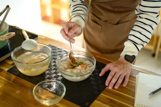 Hands seen seasoning a clear broth soup with meatballs and herbs.