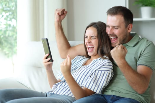 Excited couple checking mobile phone sitting in a sofa at home