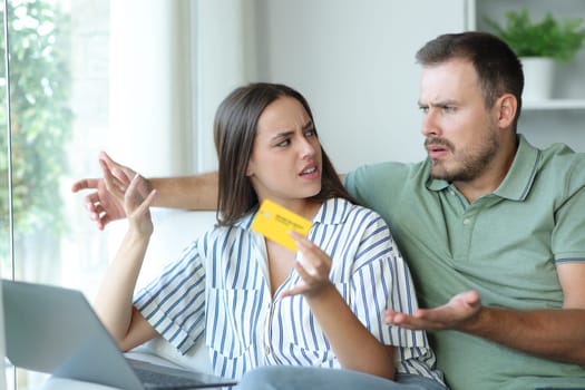 Angry couple arguing buying online with laptop and credit card sitting on a couch at home