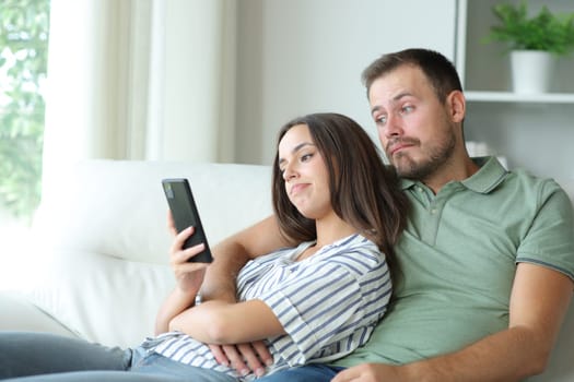 Disappointed couple checking boring phone online content sitting on a couch at home