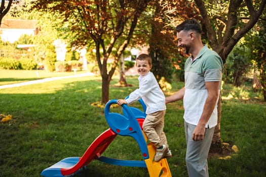 Father helps son play on a colorful slide outdoors.