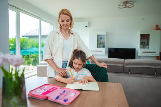 A mother helps her daughter focus on homework at a tidy desk.