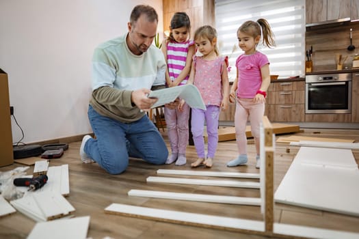 A father kneels on the floor, carefully reading instructions as his two daughters curiously observe him. The warm, inviting kitchen setting adds to the family bonding moment, filled with joy.
