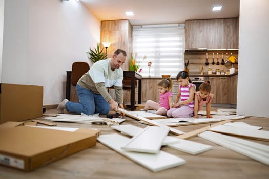 In a warm and inviting living area, a father carefully assembles furniture while surrounded by his three engaged daughters, creating a fun and collaborative home project atmosphere.