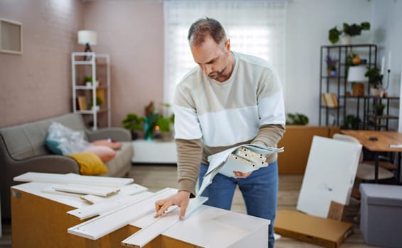 A man focuses intently on assembling furniture in a bright living room. Surrounded by parts and tools, he references instructions while creating a welcoming space filled with greenery and warmth.