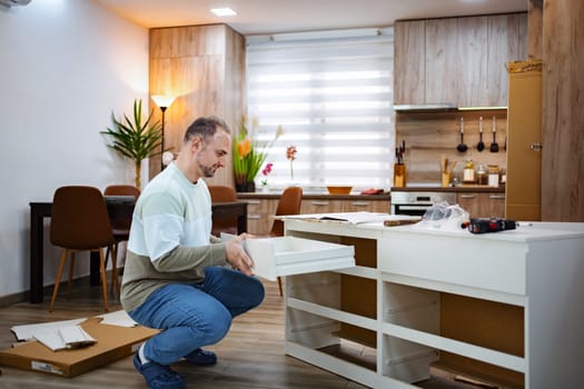 In a stylish kitchen with wooden accents, a man kneels, carefully installing a drawer into a sleek white cabinet. Sunlight filters through sheer curtains, enhancing the warm atmosphere.