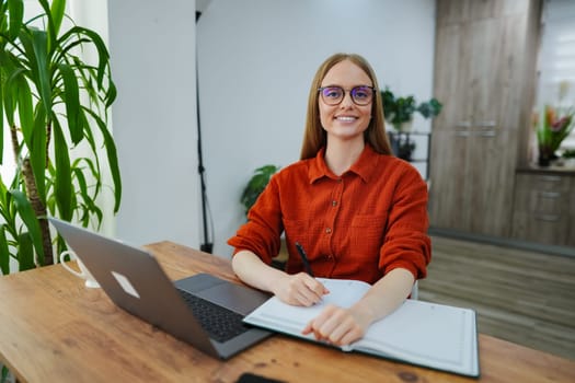 A focused woman takes notes while sitting at her desk, surrounded by greenery.