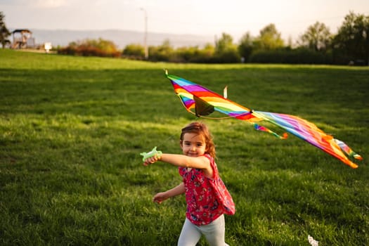 A cheerful young girl runs across the grassy field, holding a vibrant kite that dances in the gentle breeze. The warm sunlight bathes the scene, creating a joyful atmosphere of play.