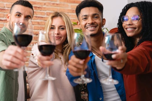 Smiling multiethnic friends raise glasses of red wine in a toast. Indoor setting with exposed brick wall, joyful atmosphere, casual outfits, and strong sense of celebration and friendship.