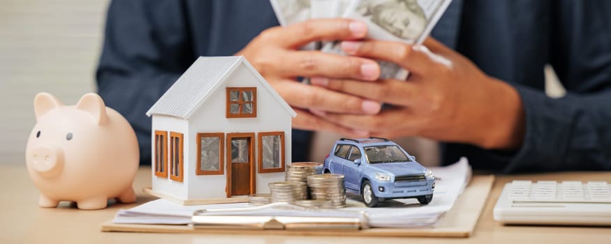 A man holding money with house and car models nearby. Highlights financial and property planning, saving cash, managing loans, and tax strategies for purchasing assets.