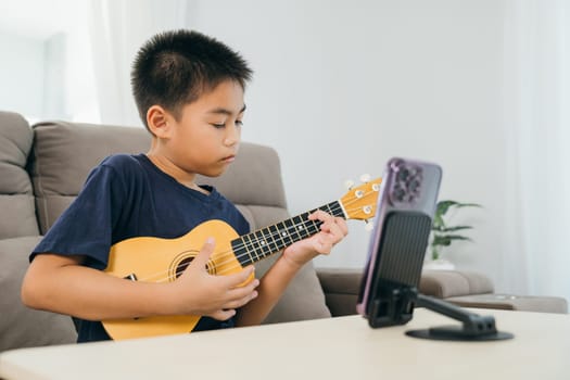 Asian kid boy playing ukulele while live-streaming his music lesson using a smartphone. The mounted phone stand and relaxed living room scene showcase modern learning and creative activities at home.
