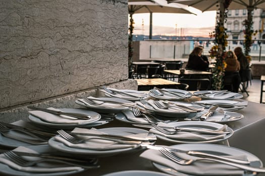 Plates neatly arranged with forks, knives, and napkins on stone countertop at outdoor restaurant in selective focus. Blurred background with European city square at sunset. Relaxed dining, people seated, umbrellas, travel destination, summer vacation