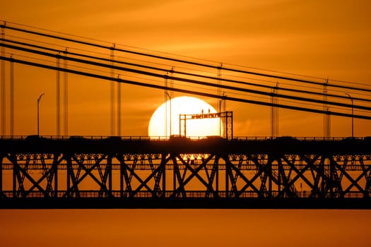 Close view of sun setting directly behind 25 de Abril bridge in Lisbon, Portugal, warm orange sky with some clouds, cars and cables in silhouette against glowing sun, striking urban sunset scene ,vehicles crossing bridge, summer heat haze, bright sun