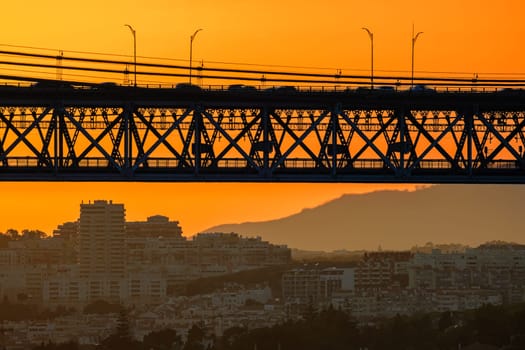 Dramatic close view of section of iconic 25 de Abril bridge in silhouette against vibrant orange sunset sky, Lisbon, Portugal cityscape and distant hills below in summer sundown haze in background, vehicles run on bridge in motion blur