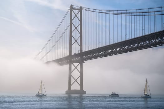 Dramatic view of Lisbon iconic 25 de Abril bridge in morning fog on sunny day, with sailboats and motorboat on tranquil Tagus river, Portugal. Misty atmosphere, serene, mysterious scene, perfect for travel, cityscape themes, suspension bridge.
