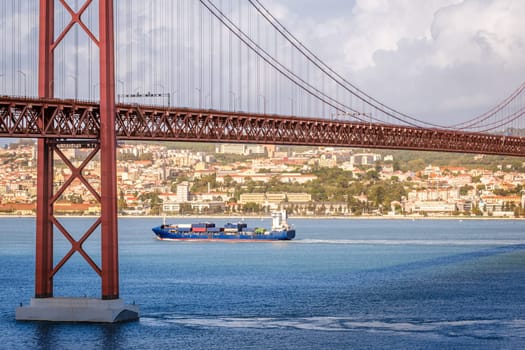 Colorful landscape with blue container ship on Tagus river under iconic 25 de Abril or 25th April bridge, Lisbon, Portugal. Famous ironwork of red suspension bridge, vibrant cityscape, urban architecture, maritime transport, sunny day, cloudy sky