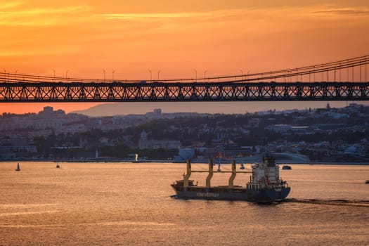 Cargo ship sail on Tagus river under iconic 25 de Abril bridge in silhouette, Lisbon, Portugal at sunset. City skyline, glowing orange sky, dramatic maritime and urban scene, European port, summer haze over horizon, tiny sailboats on calm waters.