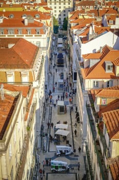 Overhead perspective of busy pedestrian street lined with historic buildings and red-tiled rooftops, people walking, touristic tuk-tuks, traditional pavement and outdoor cafes, vibrant atmosphere of city downtown, Lisbon, Portugal, on sunny day.
